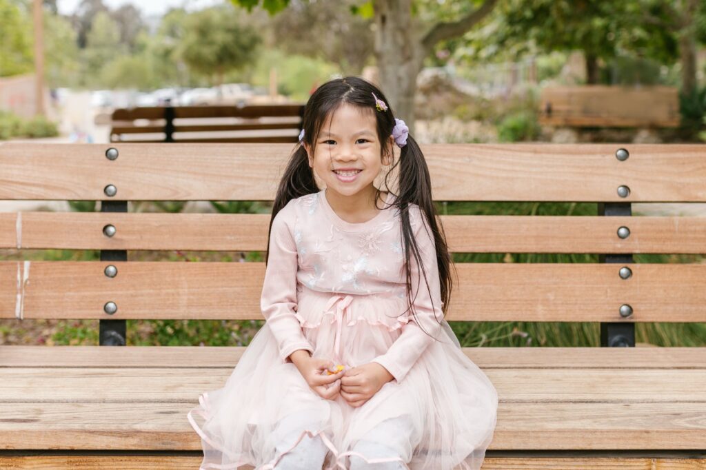 a young girl in pink dress smiling while sitting on a wooden bench