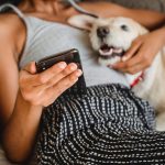 crop ethnic woman with smartphone embracing cute dog on bed